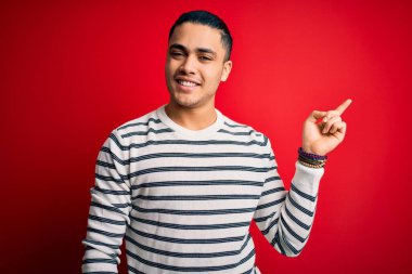 İzole edilmiş kırmızı arka planda duran sıradan çizgili tişört giyen genç Brezilyalı bir adam yüzünde büyük bir gülümsemeyle kameraya el ile işaret ediyor..