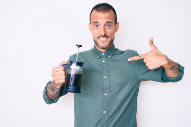 Dövmeli genç ve yakışıklı bir adam, elinde Fransız kahve makinesi olan mutlu ve gururlu bir kişiyi işaret ediyor. 