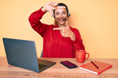 Çağrı merkezinde operatör kulaklığı takan genç esmer kadın gülümsüyor. Elleriyle ve parmaklarıyla gülen yüzüyle çerçeve yapıyor. yaratıcılık ve fotoğrafçılık kavramı. 