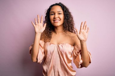 Kıvırcık saçlı, rahat tişörtlü, pembe arka planda duran ve on numarayı gösteren genç ve güzel bir kadın. Kendinden emin ve mutlu bir şekilde gülümsüyor..