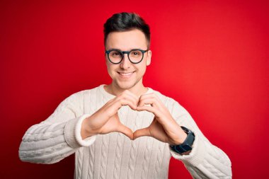 Gözlük takan yakışıklı beyaz bir adam ve kırmızı arka planda gelişigüzel bir kazak giyiyor. Aşkla gülümsüyor. Elleriyle kalp sembolü ve şekli gösteriyor. Romantik konsept.