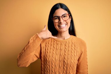 Sarı izole edilmiş arka planda gözlük takan genç, İspanyol bir kadın telefonla el ve parmak hareketleriyle gülümsüyor, telefonda konuşuyor gibi. İletişim kavramları.