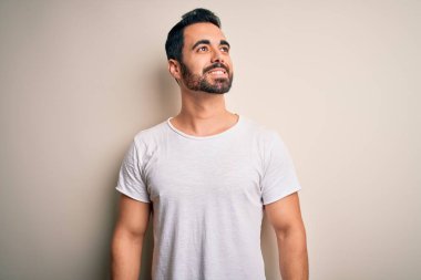 Sakallı, sıradan tişörtlü, beyaz arka planda duran, yüzünde gülümseme, doğal bir ifade olan genç yakışıklı bir adam. Kendinden emin gülen.