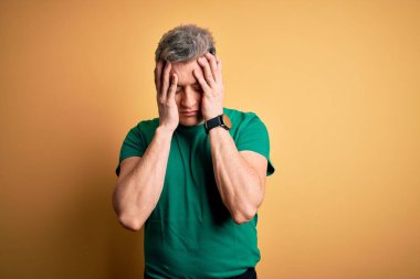 Sarı arka plan üzerine gündelik yeşil tişört giyen genç, yakışıklı, çaresiz ve stresli bir baş ağrısı çeken bir adam. Çünkü acı ve migren. Ellerini başının üstüne koy..