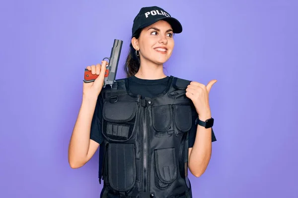 Young police woman wearing security bulletproof vest uniform and holding gun pointing and showing with thumb up to the side with happy face smiling