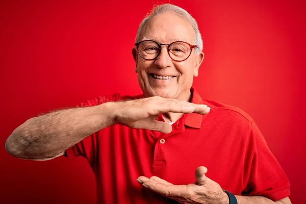 Grey haired senior man wearing glasses and casual t-shirt over red background gesturing with hands showing big and large size sign, measure symbol. Smiling looking at the camera. Measuring concept.