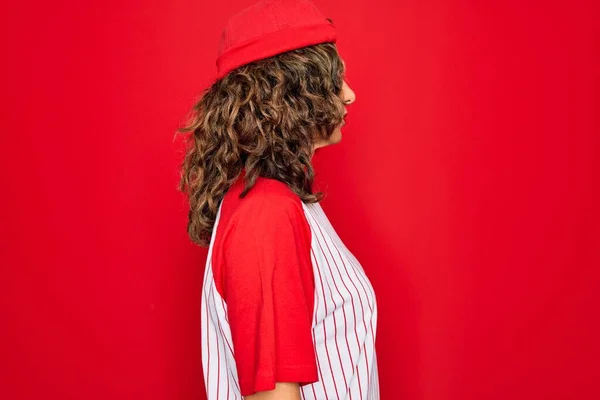 中年シニア女性身に着けている野球用具上の赤い隔離された背景を見ます側 リラックスプロファイルポーズとともに自然な顔とともに自信に満ちた笑顔 — ストック写真