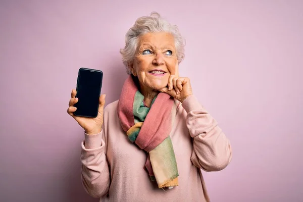 ピンクの背景の上にスマートフォンの画面を保持しているシニア美しい白髪の女性深刻な顔の質問について考えて 非常に混乱したアイデア — ストック写真