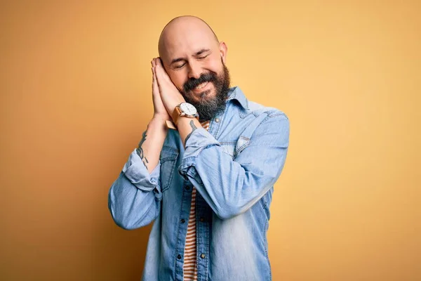 Handsome bald man with beard wearing casual denim jacket and striped t-shirt sleeping tired dreaming and posing with hands together while smiling with closed eyes.