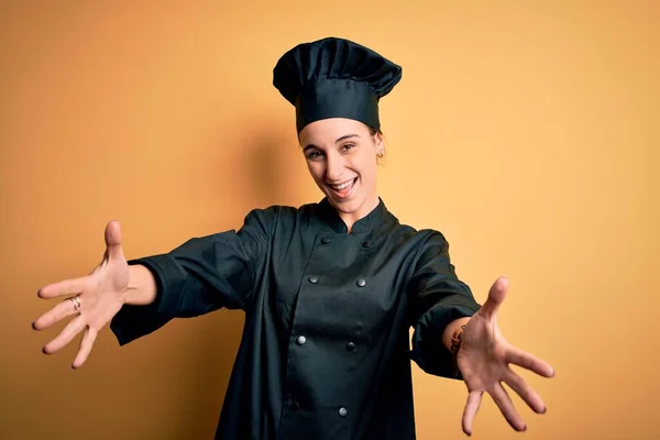 炊飯器の制服を着た若い美しいシェフの女性と抱擁のための開いた腕で笑顔カメラを見て黄色の背景に立って帽子 幸せを受け入れる陽気な表情 — ストック写真