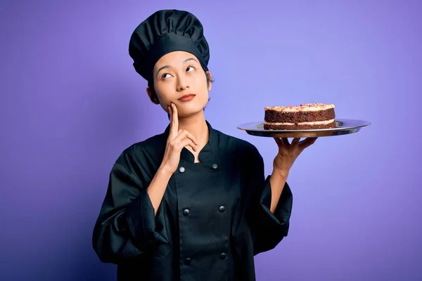 年轻的中国厨师女人穿着炊事服 拿着蛋糕盘 脸上挂着严肃的表情 思考着问题 非常迷惑的想法 — 图库照片