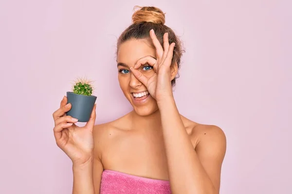 Beautiful blonde woman with blue eyes wearing towel shower after bath holding small cactus with happy face smiling doing ok sign with hand on eye looking through fingers