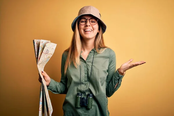 Blonde explorer woman with blue eyes on vacation wearing hat and binoculars holding city map very happy and excited, winner expression celebrating victory screaming with big smile and raised hands