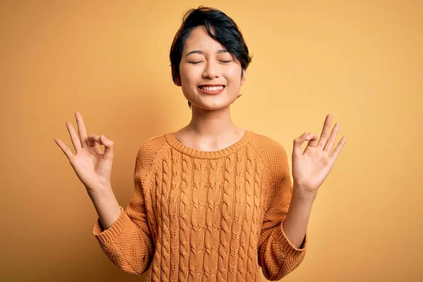 カジュアルなセーターを着た若い美しいアジアの女の子と黄色の背景の上に立っているダイエムリラックスし 指で瞑想ジェスチャーを行う目を閉じて笑顔 ヨガのコンセプト — ストック写真