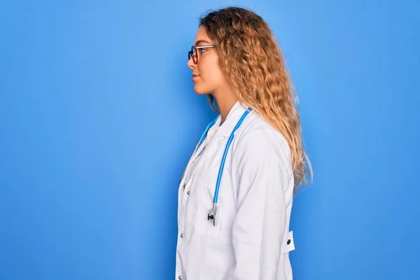 Young beautiful blonde doctor woman with blue eyes wearing coat and stethoscope looking to side, relax profile pose with natural face with confident smile.