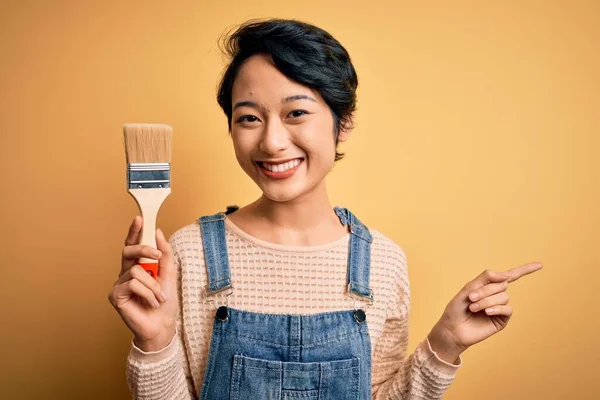 Young beautiful chinese woman painting holding paint brush over isolated yellow background very happy pointing with hand and finger to the side