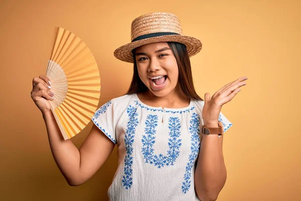 年轻的亚洲旅游姑娘戴着夏帽 手执扇子披着黄色的背景 非常高兴而兴奋 获胜者的表情洋洋得意地欢呼胜利 举起双手庆祝胜利 — 图库照片