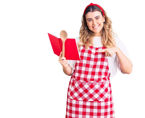 Νεαρή Καυκάσια Γυναίκα Φορώντας Ποδιά Κρατώντας Βιβλίο Συνταγών Και Κουτάλι — Φωτογραφία Αρχείου