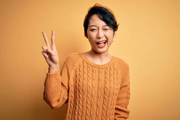 Young beautiful asian girl wearing casual sweater and diadem standing over yellow background smiling with happy face winking at the camera doing victory sign with fingers. Number two.