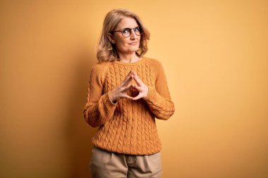 Sarışın, orta yaşlı, güzel bir kadın sarı arka planda gözlük takıyor eller yan yana, gülümseyerek gevşemiş ve neşeli. Başarı ve iyimser