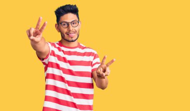 Yakışıklı Latin Amerikalı, sıradan kıyafetler ve gözlük takan, ağzı açık gülümseyen, iki elini de zafer işareti yapan genç adam. İki numara.. 