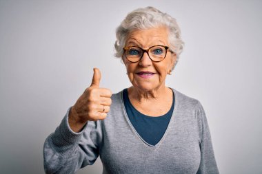 Son sınıf güzeli, gri saçlı, sıradan süveter giyen ve beyaz arka planda gözlük takan güzel bir kadın el hareketleriyle mutlu mesut el hareketi yapıyor. Kameraya bakıp başarıyı gösteren ifade onaylanıyor.