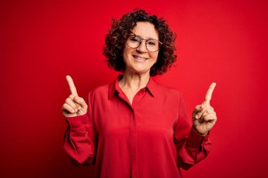 Orta yaşlı güzel kıvırcık saçlı kadın kırmızı arka planda gözlük takıyor ve farklı yönleri gösteren kendine güvenle gülümsüyor. Reklam için boşluk kopyala