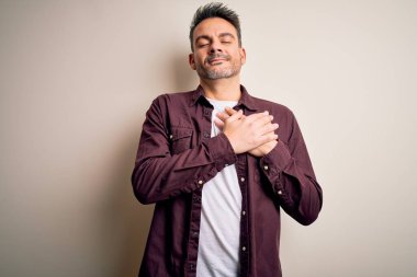 İzole edilmiş beyaz arka plan üzerinde duran, elleri göğsünde, gözleri kapalı ve yüzünde minnet dolu bir jest olan genç, yakışıklı bir adam. Sağlık kavramı.