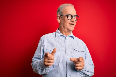 Orta yaşlı, beyaz, sıradan çizgili gömlek giyen ve kırmızı arka planda gözlük takan yakışıklı adam mutlu ve komik yüzlü parmaklarını kameraya doğrultuyor. İyi enerji ve titreşimler.