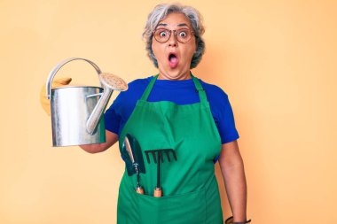 Bahçıvan önlüğü ve eldiven giyen yaşlı, beyaz saçlı bir kadın. Elinde su kovası olan bir eldiven. Şaşırtıcı bir şekilde ağzı açık ve korkmuş. 