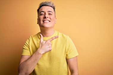 Sarı izole edilmiş arka plan üzerinde sarı tişört giyen genç ve modern bir adam. Yüzünde mutlu ve doğal bir ifadeyle el ve parmak ile işaret eden bir gülümseme var.