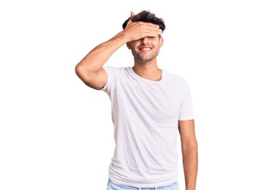 Gündelik kıyafetler giyen, gülümseyen ve gülümseyen genç İspanyol adam şaşırmak için gözlerini kapatıyor. kör kavram. 