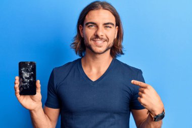 Kırık akıllı telefon tutan genç yakışıklı adam kırık ekranı gösteriyor mutlu ve gururlu gülümseyen bir kendini işaret ediyor. 