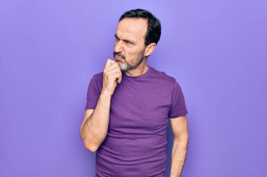 Orta yaşlı, mor tişört giyen yakışıklı bir adam izole edilmiş arka planda dikilirken parmak çenesinde şüpheye yoğunlaşır ve yukarı bakar.