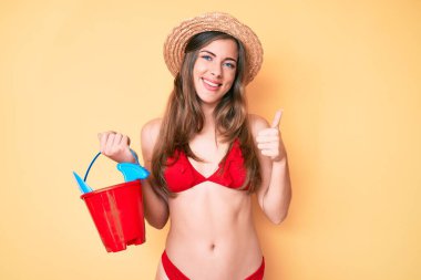 Güzel beyaz kadın yaz küreği ve kova oyuncaklarıyla oynuyor mutlu ve pozitif gülümsüyor, başparmağıyla mükemmel ve onay işareti yapıyor. 