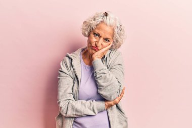 Yaşlı, gri saçlı, günlük spor giysiler giyen, kollarını kavuşturup depresyondan sıkılmış ve yorgun görünen bir kadın.. 