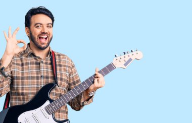 Elektro gitar çalan genç İspanyol adam parmaklarıyla imza atıyor, gülümseyerek mükemmel bir sembol oluşturuyor. 