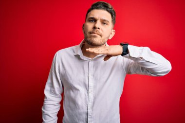 Mavi gözlü, zarif gömlekli genç bir iş adamı kırmızı izole edilmiş arka planda duruyor bıçakla boğazı kesiyor, saldırganlığı şiddetli bir şiddetle tehdit ediyor.