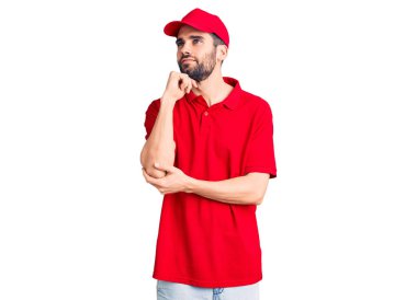 Sakallı, teslimat üniforması giyen, elleri çenesinde soru soran, düşünceli bir genç adam. Gülümseyen ve düşünceli bir yüz. şüphe kavramı. 