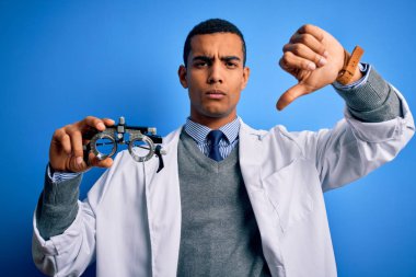 Yakışıklı afro-amerikan optik adam elinde optometri gözlüklerini mavi arka planda tutuyor, kızgın yüzlü, negatif işaretli başparmakları aşağıda, reddedilme konsepti