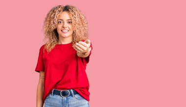 Kıvırcık saçlı, sıradan kırmızı tişörtlü genç bir kadın dostça bir selamlama ve hoş geldin hediyesi olarak gülümsüyor. başarılı iş. 