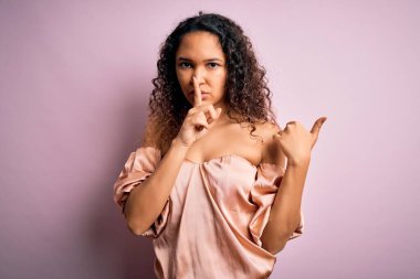 Kıvırcık saçlı, pembe arka planda sıradan tişörtler giyen genç ve güzel bir kadın ellerini yan tarafa doğru gösteren dudaklarıyla sessiz olmak istiyor. Sessizlik ve gizli konsept.