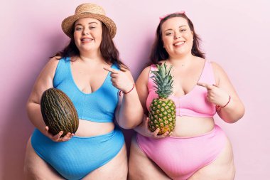Bikini giyen, kavun tutan, ananas tutan ve mutlu gülümseyen genç ve büyük beden ikizler. 