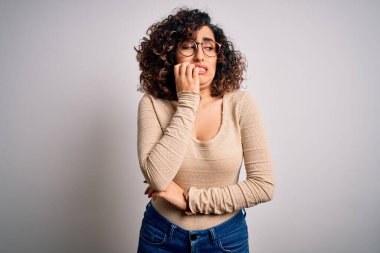 Genç, kıvırcık, kıvırcık, rahat tişört giyen ve beyaz arka planda gözlük takan genç, gergin ve dudakları tırnak yiyen bir kadın. Endişe sorunu.