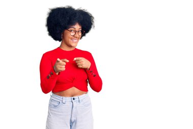 Sıradan kıyafetler giyen ve gözlüklü genç Afro-Amerikan kızı mutlu ve komik yüzüyle kamerayı işaret ediyor. iyi enerji ve titreşimler. 