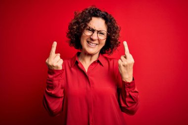 Middle age beautiful curly hair woman wearing casual shirt and glasses over red background Showing middle finger doing fuck you bad expression, provocation and rude attitude. Screaming excited