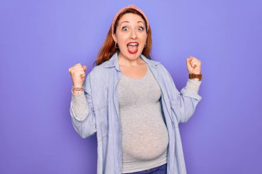 Güzel, kızıl saçlı hamile bir kadın mor arka planda bebek bekliyor. Kollarını kaldırıp, gözlerini açarak başarıyı kutluyor. Kazanan konsept.