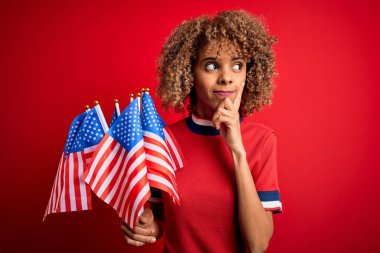 Genç Afro-Amerikan vatansever kıvırcık kadın kırmızı arka planda Birleşik Devletler bayraklarını tutuyor.