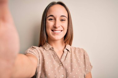 Yazlık tişört giyen genç bir kadın beyaz arka planda kamerayla selfie çekiyor. Mutlu bir yüzü var. Kendine güvenen bir gülümsemeyle dişlerini gösteriyor.