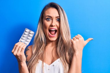 Sarışın, güzel bir kadın ilaç haplarını mavi arka planda tutuyor. Parmağını yan tarafa uzatıyor, ağzı açık gülümsüyor.
